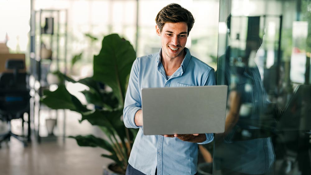 Mann in blauem Hemd steht in einem Büro und hält lächelnd einen Laptop in der Hand