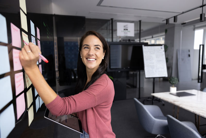 Optimierung von Arbeitsabläufen mit agilen Methoden. Selbstbewusste junge Product Ownerin organisiert den Arbeitsprozess, verfolgt Aufgaben auf einem transparenten Scrum-Board und macht sich Notizen vom Tablet, während sie lächelnd zur Seite blickt.